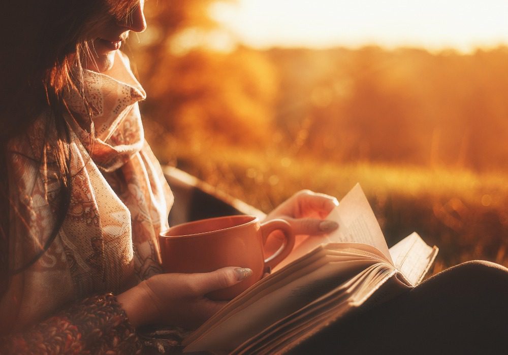 Woman in scarf reading a novel with cup in hand outside and sunset in background.