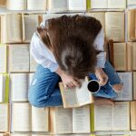 Woman sitting on a floor covered with open books, holding a black coffee and reading a book, tracing words with her finger.